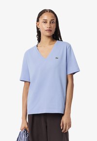 Donna con capelli intrecciati che indossa una maglietta V-neck di Lacoste blu chiaro e pantaloni scuri, teneva un oggetto di stoffa a righe nella mano sinistra.