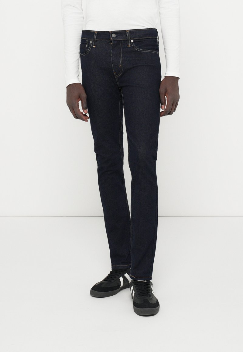 Jean en denim bleu foncé avec une coupe slim, design à cinq poches, coutures contrastantes et fermeture à bouton métallique, assortis à des baskets noires.