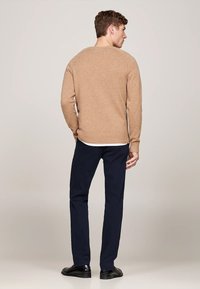 Maglione lavorato a maglia color sabbia con scollatura rotonda e orlo a coste, abbinato a pantaloni blu scuro e scarpe nere, visto da dietro.