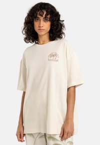 Lys beige oversized T-skjorte laget av bomull, med et palme-tre grafikk i jordtoner på brystet og rund halslinning.