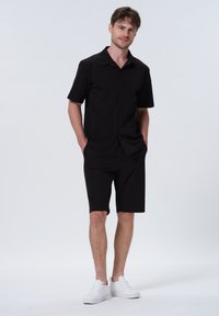 Schwarzes Kurzarm-Hemd mit Knopfverschluss, kombiniert mit passenden knielangen Shorts. Das Outfit hat einen lässigen Schnitt und wird von weißen Sneakers ergänzt.