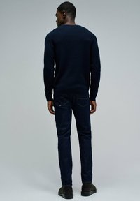 Hombre de pie de espaldas, vistiendo un suéter azul marino oscuro, jeans azul oscuro y botas negras, contra un fondo gris liso.