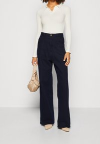 Nainen, joka on pukeutunut ribattyyn valkoiseen pitkähiharaiseen paitaan, korkeavyötäröisiin tummansinisiin leveälahkeisiin housuihin, vaaleisiin kärjellisiin kenkiin ja pitää kädessään vaaleaa solmittua käsilaukkua.