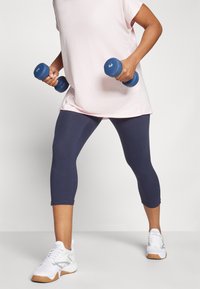 Jasnoróżowa sportowa bluzka, granatowe krótkie legginsy oraz białe trampki. Trzyma niebieskie hantle z oznaczeniem wagi. Gładka tkanina, luźny krój.