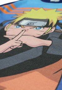 Tissu bleu présentant un design d'un personnage aux cheveux blonds ébouriffés avec un geste de la main. Des accents gris et orange rehaussent le graphisme.