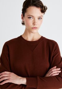 Sudadera marrón de cuello redondo con un acabado texturizado, presenta puños y cuello acanalados. El modelo está de pie con los brazos cruzados, mostrando el ajuste de la prenda.