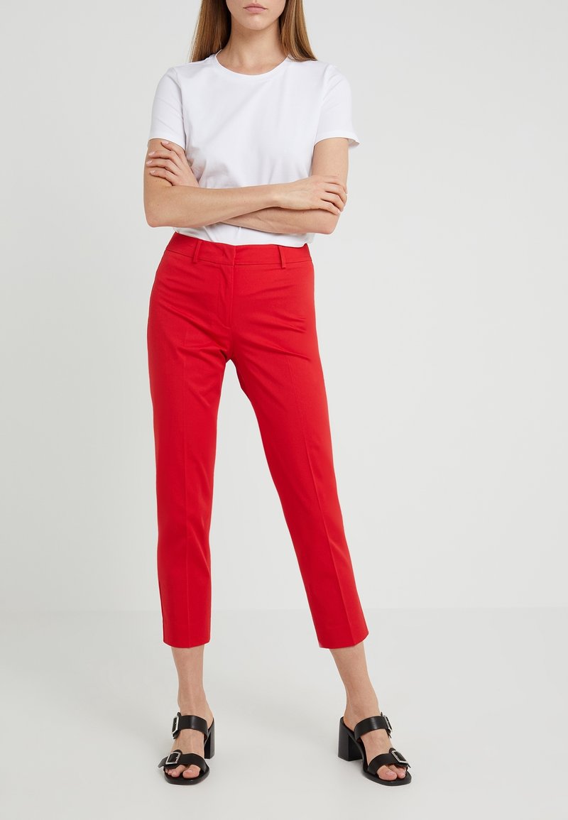 Personne portant un pantalon rouge court, une chemise blanche à manches courtes et des sandales à talons noires, debout, les bras croisés.