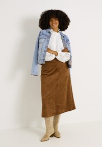 Jupe maxi en daim marron assortie à une veste en denim bleu clair, une chemise blanche avec une finition en dentelle et des bottes de cowboy beige clair.