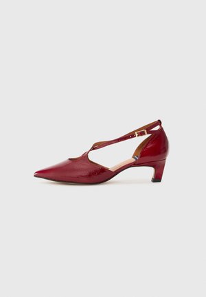Red patent leather pump with a pointed toe, cut-out sides, and a mid-heeled design. Features an adjustable strap for secure fit.