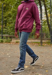 Hoodie bordeaux avec logo, poche avant et cordon de serrage. Jean bleu délavé à coupe ajustée. Baskets noires et blanches avec surface texturée.