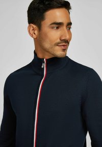 Marinekleurige zip-up trui met een hoge kraag, voorzien van rode en witte strepen langs de rits. Gebreide textuur met een getailleerd ontwerp.