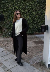 Vrouw met zwarte zonnebril, lange zwarte jas, witte trui en zwarte broek, lopend op een bestrate weg naast een muur bedekt met klimop.