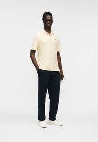 Κοντομάνικο φωτεινό κίτρινο πόλο πουκάμισο με διακριτικές ρίγες, συνδυασμένο με σκούρο μπλε παντελόνι και λευκά αθλητικά παπούτσια. Απλός, μοντέρνος σχεδιασμός.