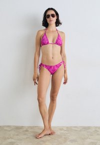 Rožinis raštuotas bikini maudymosi kostiumėlis su trikampiu viršumi ir nugaros raiščio apatine dalimi. Modelis dėvi akinius nuo saulės. Lygus paviršius, minimalistinis dizainas ir ryški spalva.