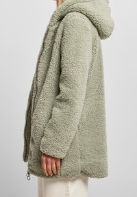 Chaqueta de forro polar en verde claro, con capucha, bolsillos laterales y una superficie texturizada. Cierre de cremallera con un acento en forma de anillo.