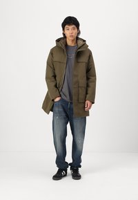 Veste imperméable vert olive avec capuche, poches avant et coupe décontractée ; portée sur un t-shirt gris et un jean bleu déchiré. Baskets noires.