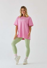 Przesadzona różowa bawełniana koszulka z napisem "Pilates Princess", zestawiona z jasnoskalnymi legginsami i białymi sneakersami w różowo-czerwone paski.