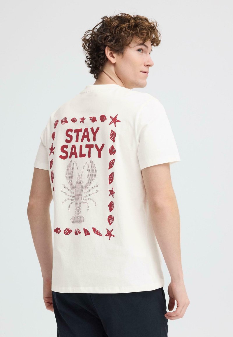 Mladić s kovrčavom kosom nosi bijelu majicu na kojoj se nalaze crveni jastog, školjke, morska zvijezda i tekst "OSTANI SLAN" na stražnjoj strani.