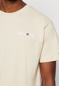 Beige cotton t-shirt with a round neck, featuring white embroidered "Tommy Jeans" logo on the left chest and a small logo detail on the sleeve.