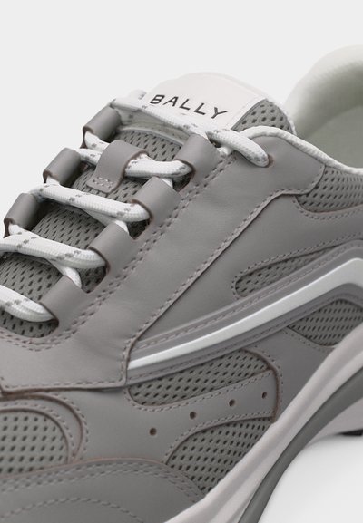 Κοντινή λήψη ενός γκρι και λευκού αθλητικού παπουτσιού Bally με πάνελ από δίχτυ και δέρμα και λευκά κορδόνια που περνούν μέσα από καφέ μεταλλικά κρικάκια.