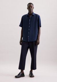 Camicia a maniche corte di colore navy, chiusa con bottoni e realizzata in tessuto leggero, abbinata a pantaloni scuri di taglio ampio e scarpe nere con lacci.