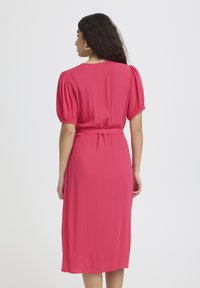 Femme aux longs cheveux foncés portant une robe rose vif à manches courtes, longueur genou, avec des manches froncées et une ceinture à la taille, vue de dos.