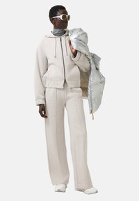 Beige, übergroßer Hoodie und passende weite Hosen aus weichem Stoff, kombiniert mit einer glänzenden silbernen Daunenjacke und weißen Sonnenbrillen.