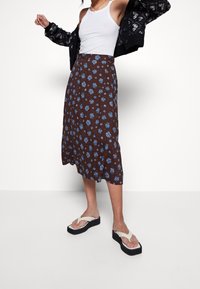 Falda midi marrón con estampado de flores azules, tela fluida, combinada con sandalias beige tipo chancla y un cárdigan negro con estampado.