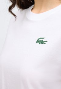 Camiseta blanca de algodón con un pequeño logo de cocodrilo verde en el pecho, cuello redondo y textura suave.