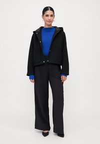 Vrouw die staat, gekleed in een zwarte capuchonjas, felblauwe trui, brede zwarte broeken en zwarte puntige schoenen, tegen een effen witte achtergrond.