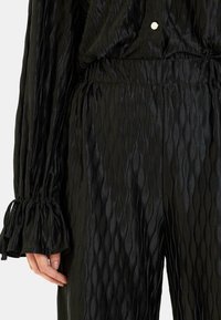Czarny top z plisowanej tkaniny z teksturowanym wzorem falistym, elastycznym paskiem w talii oraz zgromadzonymi rękawami z wiązaniami. Obecny akcent w postaci srebrnego guzika.