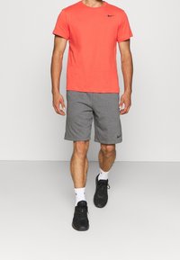 Mężczyzna w jasnopomarańczowej koszuli z krótkim rękawem, szarych krótkich spodniach do kolan, białych skarpetach sportowych i czarnych butach sportowych stoi na tle jednokolorowym.