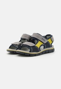 Marinblå och grå sandal med gula detaljer; tillverkad av mocka och syntetiska material; har justerbara kardborreband och en öppen tådesign.