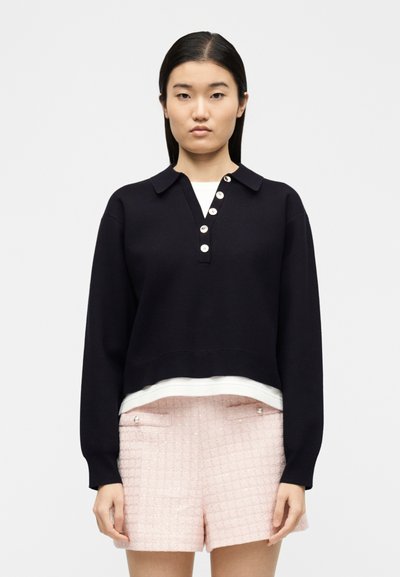 Νεαρή γυναίκα με ίσια σκούρα μαλλιά, ντυμένη με μαύρο πουλόβερ με κουμπιά και γιακά, πάνω από λευκό πουκάμισο και ροζ σορτς με υφή.