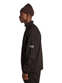 Mand iført mørkebrun hue og sort lynlåstrøje med "Halo" patch på ærmet, stående i profil mod hvid baggrund.