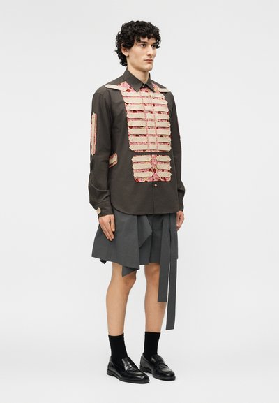 Mees, kes kannab tumedat särki beeži ja lillemustriga paneelidega, kombineerituna asümmeetrilise halli seelikuga, mustade sokkide ja mustade kingadega.