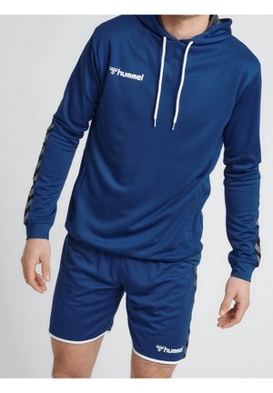 Mann trägt blaue Hummel-Sportkapuzenjacke mit weißen Kordeln und passende blaue Shorts mit Markenlogos auf Brust und Bein.
