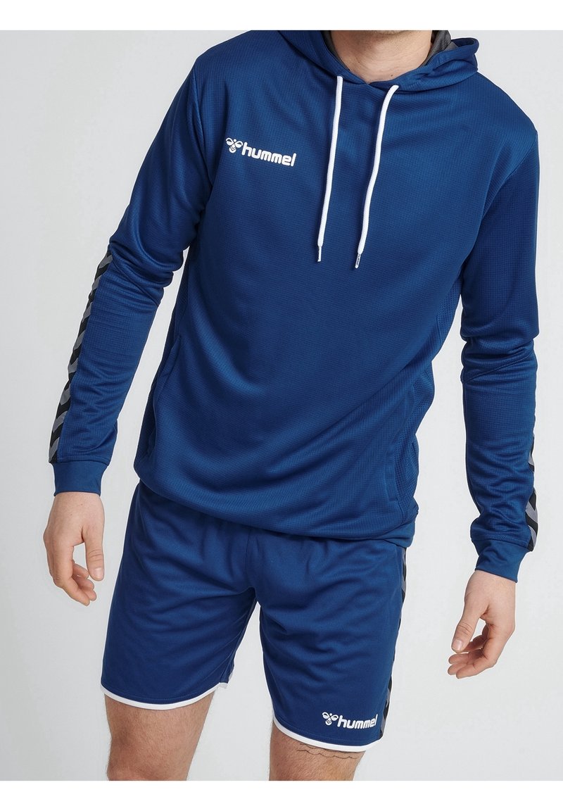 Homme portant un sweat à capuche de sport bleu hummel avec des cordons blancs et un short bleu assorti avec les logos de la marque sur la poitrine et la jambe.
