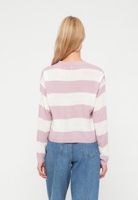 Femme aux longs cheveux blonds portant un pull à rayures horizontales larges rose et blanc, ainsi que des jeans bleus, se tenant devant un fond blanc uni.