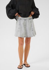 Gonna di denim grigia chiara con design svasato, caratterizzata da cuciture a contrasto e una vita piatta, abbinata a una maglietta nera larga e sandali.