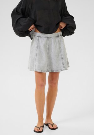 Gonna di denim grigia chiara con design svasato, caratterizzata da cuciture a contrasto e una vita piatta, abbinata a una maglietta nera larga e sandali.