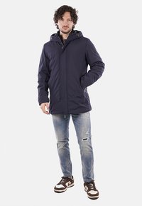 Giacca impermeabile blu navy con cappuccio, zip frontale, tasche con zip e polsini elasticizzati. Indossata sopra jeans chiari strappati e sneakers.