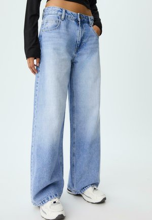 Personne portant un jean large bleu clair, des baskets blanches, et un top court noir à manches longues avec une main dans la poche.