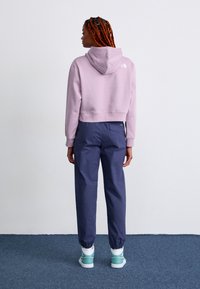 Haut à capuche en lavande coupé, avec une capuche et un logo blanc au dos, associé à un pantalon bleu marine à taille élastique et des chaussures à accents turquoise.