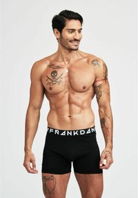 Boxer-briefs noirs avec une ceinture logo blanche, en coton, au design ajusté, texture lisse et détails de tatouages visibles sur les bras.