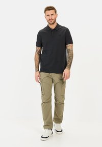 Sort polo-shirt med et diskret logo, sammen med lyse olivenfarvede cargo-bukser med flere lommer og lynlåse, kompletteret af hvide sneakers.
