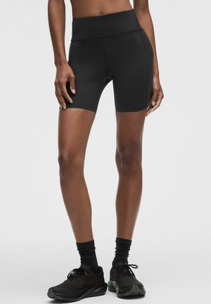 Persoon die zwarte, hoog getailleerde strakke sportshorts draagt, zwarte sokken en zwarte hardloopschoenen, staand tegen een effen lichte achtergrond.