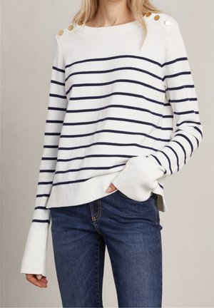 Maglione bianco a righe con strisce orizzontali blu navy, dotato di dettagli con bottoni sulle spalle, collo a coste e vestibilità rilassata. Abbinato a jeans scuri.