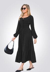 Vestido midi negro con mangas largas y cintura fruncida, que presenta un dobladillo con volantes. Combinado con zapatos negros de tacón destalonados y un bolso negro.