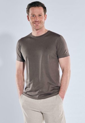 Lächelnder Mann steht mit Händen in den Taschen, trägt ein braunes T-Shirt mit kurzen Ärmeln und beige Hose vor einem schlichten hellen Hintergrund.